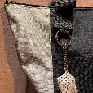 May include: A close-up of a tote bag with a black and gray color scheme. A gold keychain with a white cube-shaped charm hangs from the bag's handle. The charm has a lattice design and gold accents.