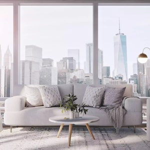 May include: A white sofa with throw pillows and a blanket sits in a living room with a view of a city skyline. A round coffee table with a plant sits in front of the sofa. A white chair with a gold floor lamp sits to the right of the sofa.