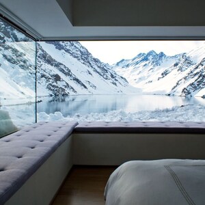 Peut inclure: Une chambre à coucher avec une grande fenêtre donnant sur une chaîne de montagnes enneigées et un lac. La fenêtre est encadrée par un mur blanc et un banc rembourré gris. Un lit blanc avec une couette blanche se trouve au premier plan.