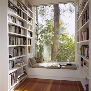 Puede incluir: Una estantería blanca empotrada con un asiento de ventana frente a una ventana grande. El asiento de ventana tiene un cojín blanco y dos almohadas. La ventana da a un árbol verde.
