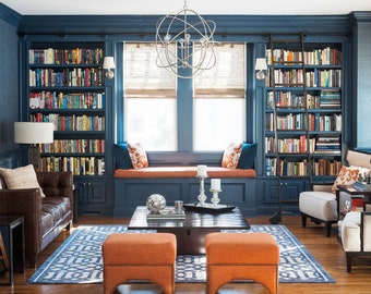 May include: A blue library with built-in bookshelves, a window seat, and two orange ottomans. The room is decorated with a chandelier, candles, and a rug.