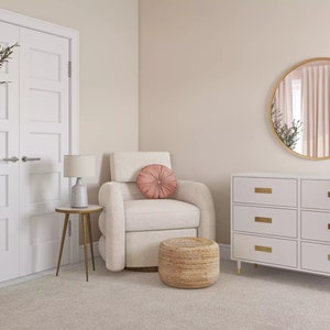 May include: A white nursery dresser with six drawers and gold hardware. A round mirror with a gold frame hangs above the dresser. A white armchair with a pink pillow sits in front of the dresser. A woven ottoman sits in front of the armchair. A potted olive tree is in the corner of the room.
