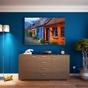 May include: A living room interior with a blue wall, a wooden dresser with drawers, two bookcases filled with books, a floor lamp with a white shade, a small toy car on the floor, and a painting of colorful houses on the wall.