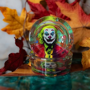 May include: A glass ashtray with a colorful image of the Joker from the movie "Joker" inside. The ashtray is sitting on a wooden surface with fall leaves around it.