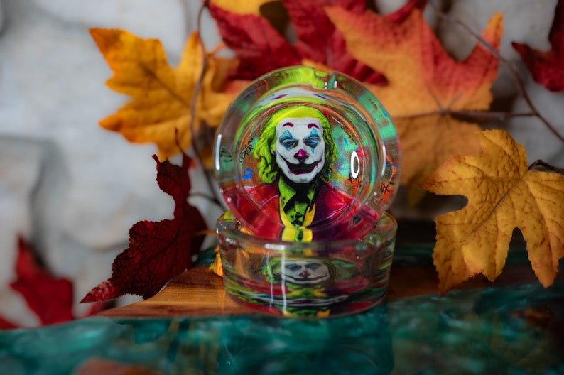 May include: A glass ashtray with a colorful image of the Joker from the movie "Joker" inside. The ashtray is sitting on a wooden surface with fall leaves around it.