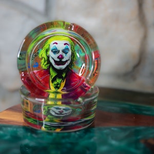 May include: A glass ashtray with a colorful image of the Joker from the movie "Joker" printed on the inside. The ashtray is stacked on top of another similar ashtray.
