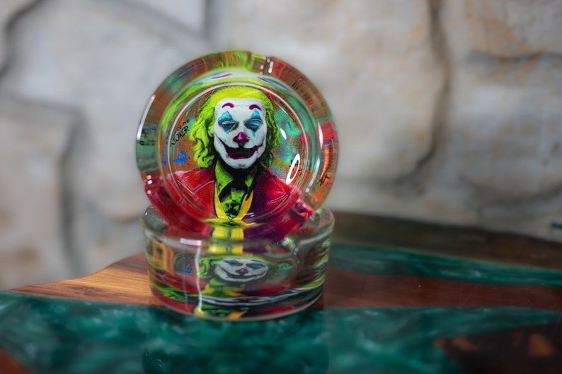 May include: A glass ashtray with a colorful image of the Joker from the movie "Joker" printed on the inside. The ashtray is stacked on top of another similar ashtray.