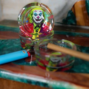 May include: Two glass ashtrays with colorful designs. The top ashtray features a Joker image. The bottom ashtray has a brown cigarette inside.