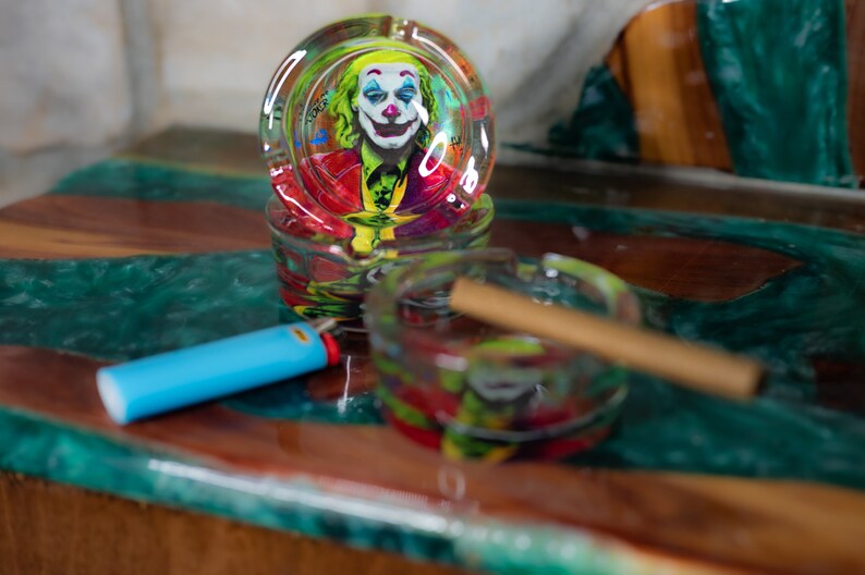 May include: Two glass ashtrays with colorful designs. The top ashtray features a Joker image. The bottom ashtray has a brown cigarette inside.