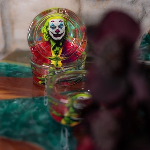 May include: A glass ashtray with a colorful image of the Joker from the movie "Joker" printed on the inside. The ashtray is sitting on a green and brown table.