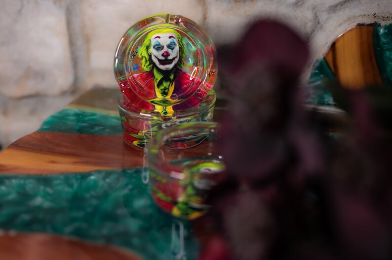 May include: A glass ashtray with a colorful image of the Joker from the movie "Joker" printed on the inside. The ashtray is sitting on a green and brown table.