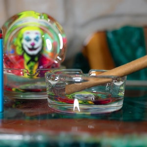 May include: Two glass ashtrays with a Joker design. One ashtray has a rolled-up cigarette inside. A blue lighter is next to the ashtrays.