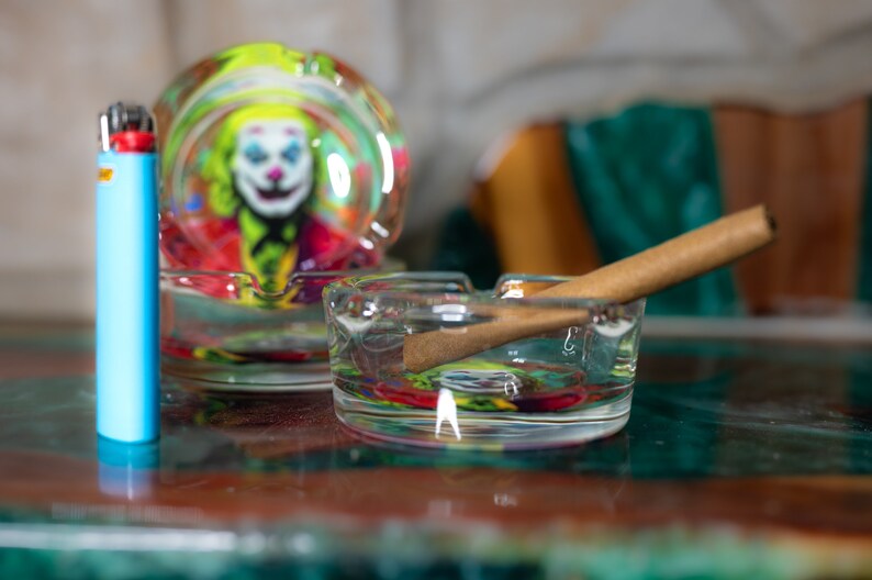May include: Two glass ashtrays with a Joker design. One ashtray has a rolled-up cigarette inside. A blue lighter is next to the ashtrays.