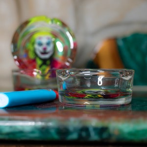May include: Two glass ashtrays with a colorful clown design. The ashtray on the left is out of focus and the ashtray on the right is in focus. A blue lighter is lying on the table in front of the ashtrays.