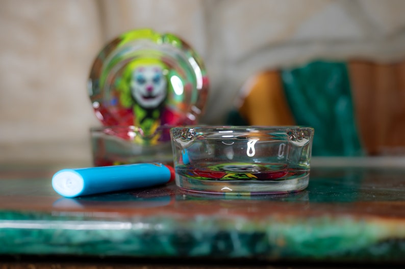 May include: Two glass ashtrays with a colorful clown design. The ashtray on the left is out of focus and the ashtray on the right is in focus. A blue lighter is lying on the table in front of the ashtrays.