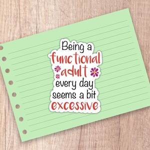May include: A sticker on a light green lined paper with the text "Being a functional adult every day seems a bit excessive" in black, red, and white. The sticker has a white border and is decorated with purple flowers.