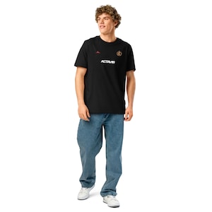 May include: A black t-shirt with white text that says "ACTAME" and a gold logo on the right side of the chest. The person wearing the shirt is standing with their legs apart and wearing blue jeans.