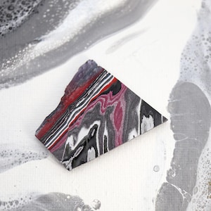 Puede incluir: Una pieza de arte triangular con un diseño veteado en tonos de rojo, negro, blanco y gris. La obra de arte tiene un borde áspero e irregular y está colocada sobre un fondo blanco y gris con un patrón fluido y abstracto.