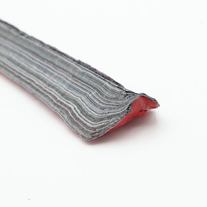 May include: A close-up of a strip of material with a layered, textured appearance. The material is primarily gray with thin white and red stripes. The bottom edge is red, and the object is set against a white background.