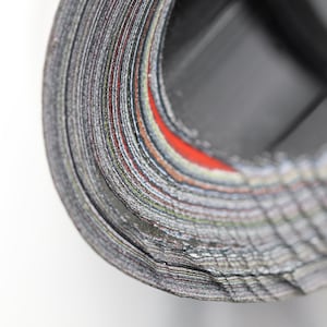 May include: Close-up of a rolled stack of paper with visible edges. The paper has a mix of gray, black, and red colors. The roll is positioned against a white background, highlighting the texture and layers of the paper.