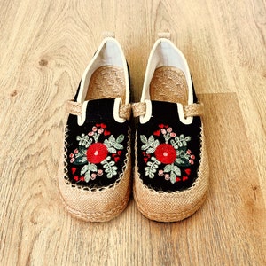 Op de afbeelding: Een paar zwarte canvas schoenen met geborduurde rode bloemen en groene bladeren. De schoenen hebben een jute zool en een witte voering.
