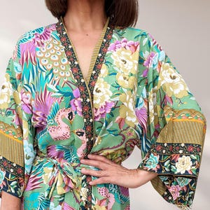 May include: A colourful floral kimono in green, pink, purple, and blue. The design features peacocks and flowers, with dark blue and gold trim on the edges and sleeves. The sleeves are wide and flowing, creating a relaxed silhouette.