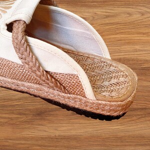 May include: Close-up of a beige espadrille sandal. The sandal has a woven jute sole, a braided strap, and a canvas upper. The design incorporates natural tones of beige and brown, with a black rubber sole. The sandal is designed for casual wear.