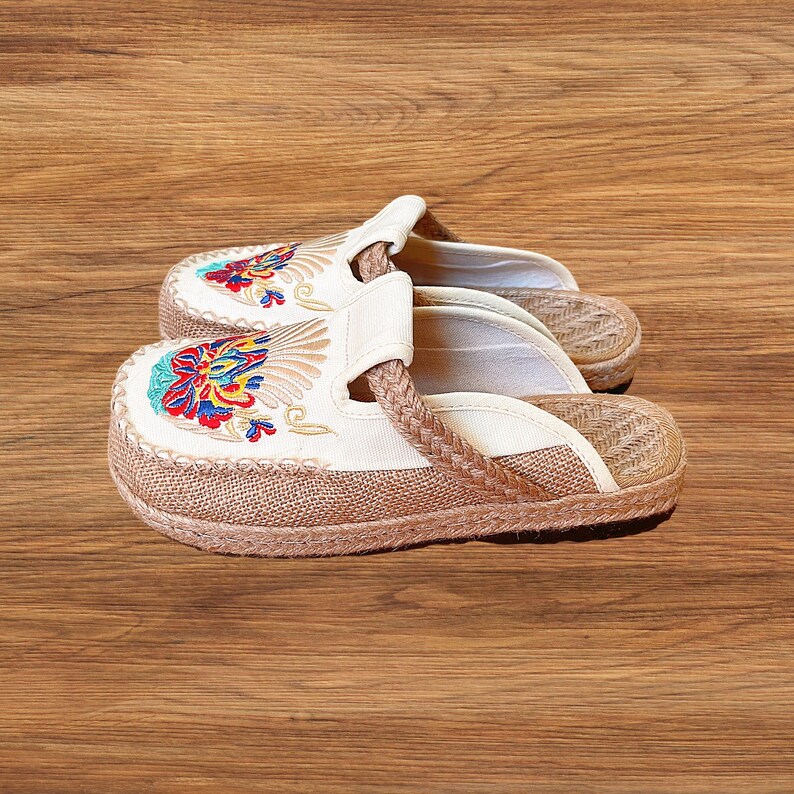 May include: Cream-coloured embroidered slippers with a woven jute sole. The slippers feature colourful floral embroidery on the toe and a braided strap. The sole is made of woven jute material.