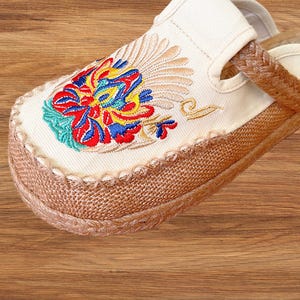 May include: Close-up of a cream slipper with a vibrant floral embroidery. The embroidery uses red, blue, yellow, and green threads. The sole is made of woven natural fibres, and the edges are stitched with cream-coloured thread.