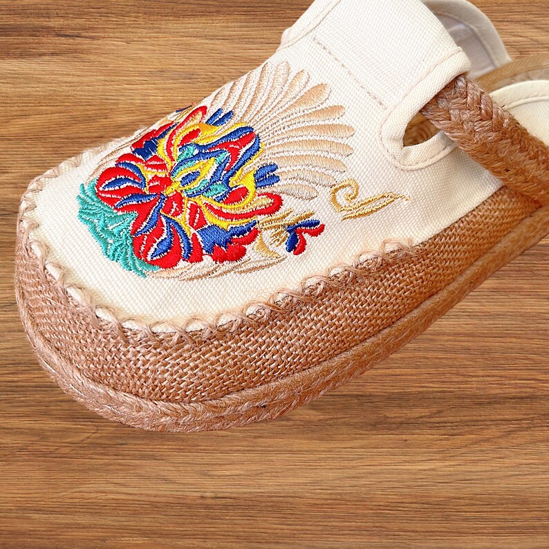 May include: Close-up of a cream slipper with a vibrant floral embroidery. The embroidery uses red, blue, yellow, and green threads. The sole is made of woven natural fibres, and the edges are stitched with cream-coloured thread.