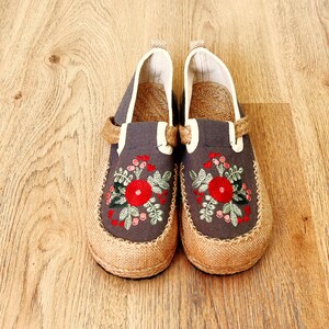 Op de afbeelding: Een paar grijze canvas slip-on schoenen met geborduurde rode bloemen en groene bladeren. De schoenen hebben een jute zool en een witte rand rond de bovenkant.