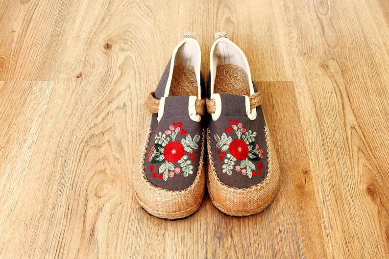 Op de afbeelding: Een paar grijze canvas slip-on schoenen met geborduurde rode bloemen en groene bladeren. De schoenen hebben een jute zool en een witte rand rond de bovenkant.