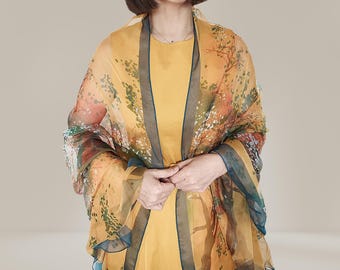 May include: A sheer, yellow shawl with a floral pattern in shades of orange, green, and brown. The shawl has a dark teal border and is draped over a yellow dress. The overall look is elegant and artistic.