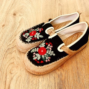Op de afbeelding: Een paar zwarte canvas slip-on schoenen met een jute zool. De schoenen zijn versierd met geborduurde rode en groene bloemen.