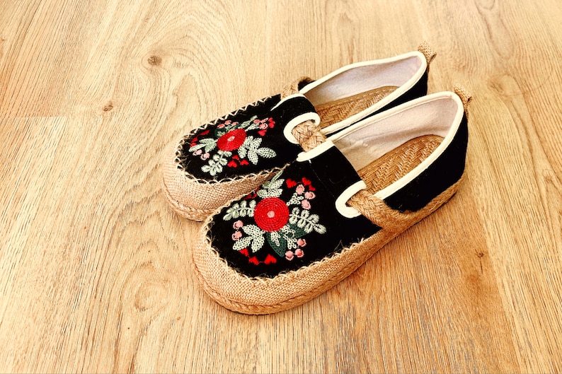 Op de afbeelding: Een paar zwarte canvas slip-on schoenen met een jute zool. De schoenen zijn versierd met geborduurde rode en groene bloemen.