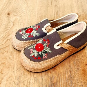 Op de afbeelding: Een paar grijze canvas slip-on schoenen met geborduurde rode bloemen en groene bladeren. De schoenen hebben een jute zool en een witte rand rond de bovenkant.
