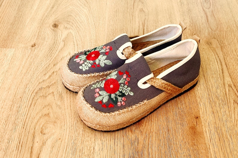 Op de afbeelding: Een paar grijze canvas slip-on schoenen met geborduurde rode bloemen en groene bladeren. De schoenen hebben een jute zool en een witte rand rond de bovenkant.