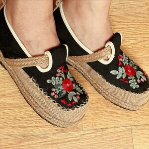 Op de afbeelding: Een paar zwarte canvas schoenen met geborduurde bloemenmotieven in rood, groen en roze. De schoenen hebben een jute zool en een witte rand rond de opening.