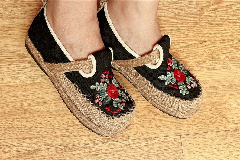 Op de afbeelding: Een paar zwarte canvas schoenen met geborduurde bloemenmotieven in rood, groen en roze. De schoenen hebben een jute zool en een witte rand rond de opening.
