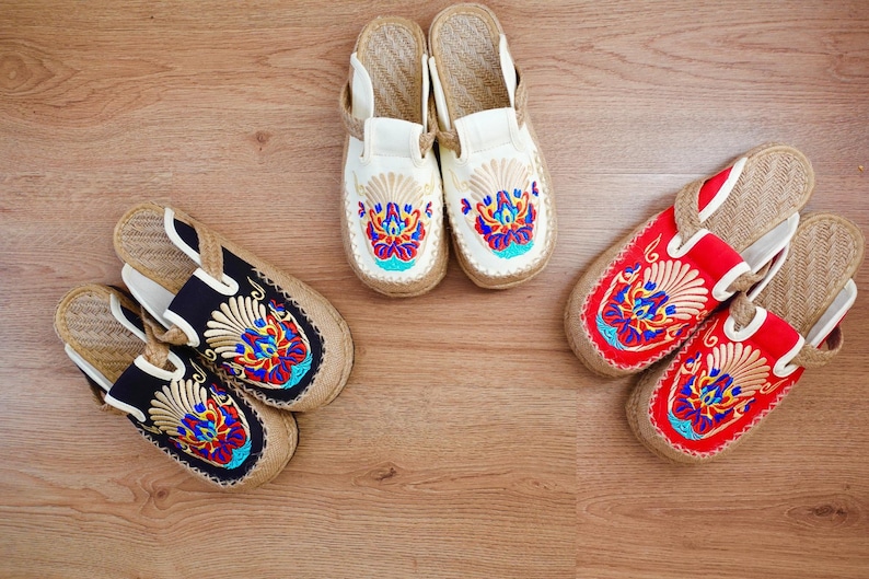 Four pairs of embroidered slippers with different coloured fabrics and designs. The slippers are made of a woven material and have a flat sole. Black, red, ivory.