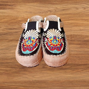 May include: Embroidered clogs with a black upper and a beige jute sole. The clogs feature a colourful floral design in red, blue, yellow, and white. The shoes have a strap for a secure fit and are set against a wooden background.
