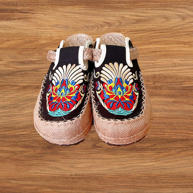 Black women’s embroidered espadrille mules, backless canvas slip on shoes, suitable for wide toe, comfortable and breathable. Embroidered clogs with a black upper and a beige jute sole. The clogs feature a colourful floral design in red, blue, yellow, and white. The shoes have a strap for a secure fit and are set against a wooden background.