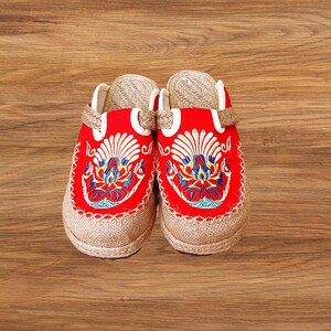 May include: A pair of red embroidered clogs with a beige jute sole. The clogs feature intricate floral embroidery in blue, yellow, and white. The upper part of the clog is red, and the sole is made of woven jute material.