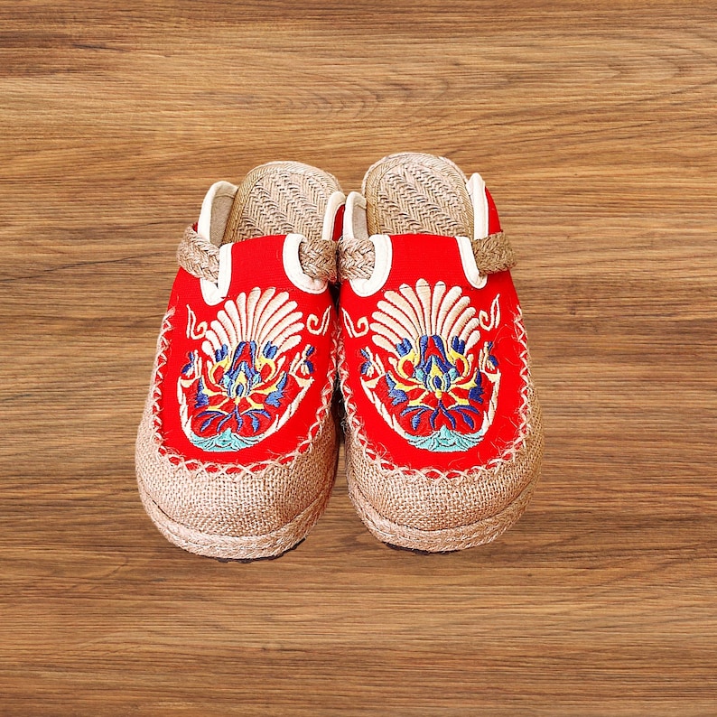 May include: A pair of red embroidered clogs with a beige jute sole. The clogs feature intricate floral embroidery in blue, yellow, and white. The upper part of the clog is red, and the sole is made of woven jute material.