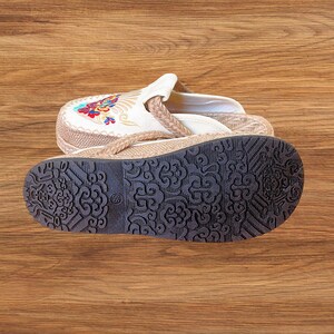 May include: A pair of beige and white embroidered sandals with a brown sole. The sandals have a floral design on the top and a braided strap. The sole is dark brown with a decorative pattern.
