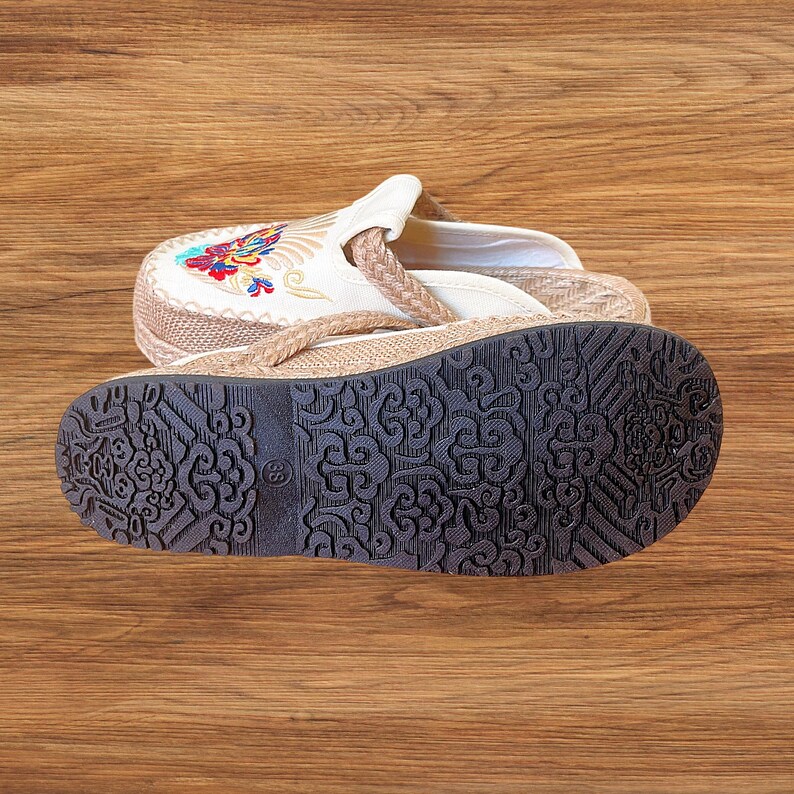 May include: A pair of beige and white embroidered sandals with a brown sole. The sandals have a floral design on the top and a braided strap. The sole is dark brown with a decorative pattern.