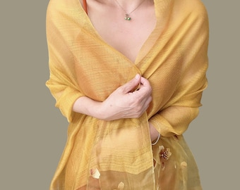 May include: A sheer, golden-yellow shawl draped over the shoulders. The fabric has a subtle texture and is adorned with a floral pattern near the bottom. A delicate necklace with a small pendant is visible.
