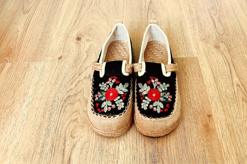 Op de afbeelding: Een paar zwarte canvas schoenen met geborduurde rode bloemen en groene bladeren. De schoenen hebben een jute zool en een witte voering.