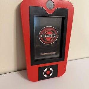 May include: A red and black handheld scanner device with a screen displaying the Chaotic logo and the text CHAOTICGAME.COM. The device features a circular button at the bottom and a circular speaker or sensor at the top, with a tapered rectangular frame.