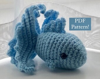 May include: A crocheted blue betta fish amigurumi toy with a long flowing tail. The toy is sitting on a white surface. PDF Pattern! is written in a white box on the image.